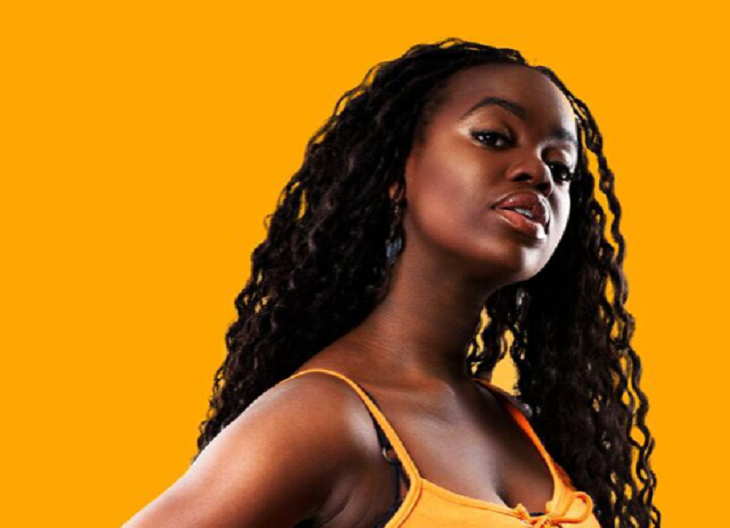 Renaida står framför en gul bakgrund. Hon har ett gult linne på sig. Hon lutar huvudet åt vänster och tittar snett in i kameran. Hon har långt svart vågigt hår.