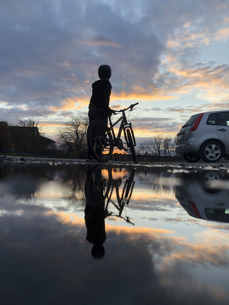 En vattenpöl i förgrunden och sedan en cyklist samt en solnedgång.